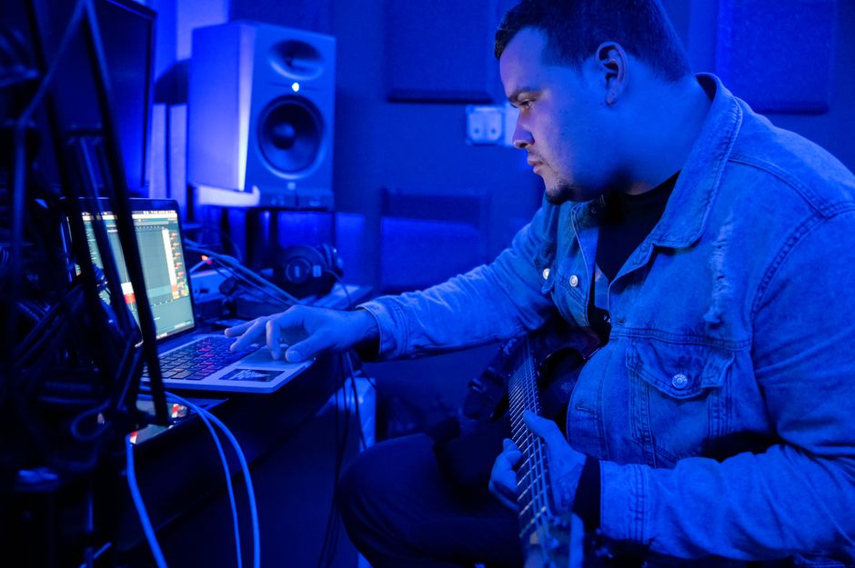 Side view of concentrated man making music on laptop and electric guitar in dark recording studio